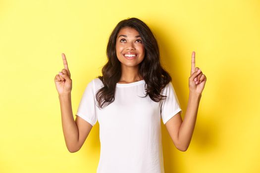 Portrait of hopeful african-american female student, looking up and smiling excited, standing over yellow background.