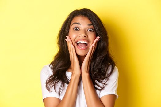 Close-up of excited, beautiful african american girl, open mouth and looking amazed at something cool, checking out an advertisement, standing over yellow background.