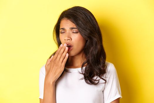 Close-up of tired and sleepy african-american girl, yawning and looking exhausted, standing in white t-shirt over yellow background.