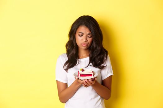 Image of sad african-american girl looking at piece of cake, cant eat it, being on diet, standing over yellow background.
