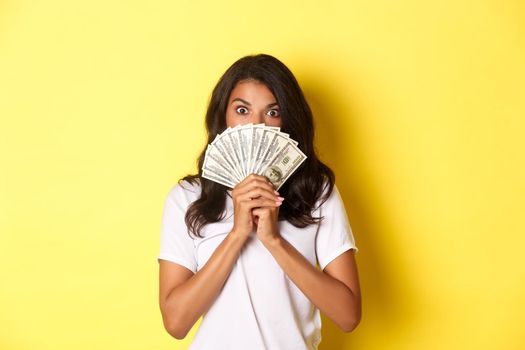Portrait of lucky african-american girl winning money prize, holding cash and looking amazed, standing over yellow background.