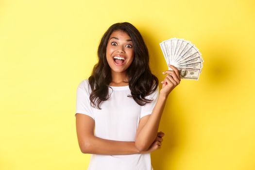 Portrait of beautiful african-american girl, smiling happy and showing money, going shopping, standing over yellow background.