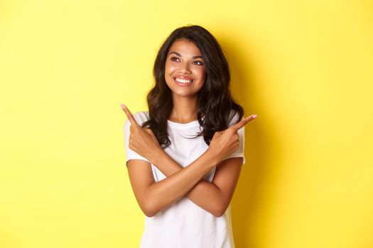 Portrait of attractive african-american girl in white t-shirt, smiling and looking left, pointing fingers sideways while making her choice between two offers, standing over yellow background.