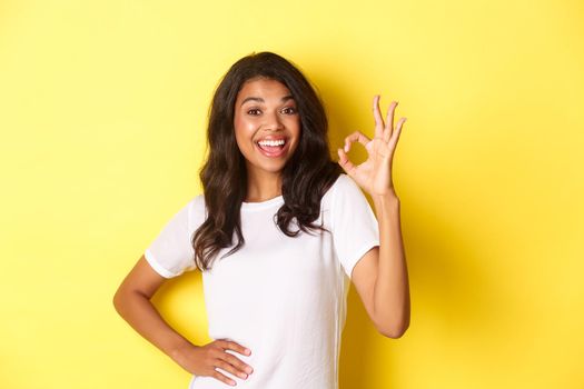 Portrait of happy good-looking african american girl, showing okay sign and smiling in approval, praise good work, standing over yellow background.
