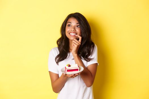 Dreamy african-american woman, smiling and looking at upper left corner thoughtful, holding piece of cake, standing over yellow background.