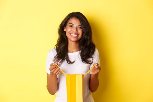 Portrait of happy and delighted african-american girl, open a shopping bad and smiling pleased, thanking for gift on holiday, standing over yellow background.