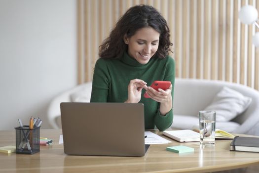 Young happy italian woman freelancer sitting at her workplace with laptop at home and using mobile phone, reading online news or chatting with friends on smartphone, watching funny videos at work
