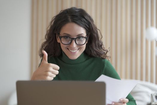 Happy young Italian female employee in glasses talks online on laptop with colleagues or business partner, showing thumb up, cheerful Spanish woman freelancer making video call while working remotely