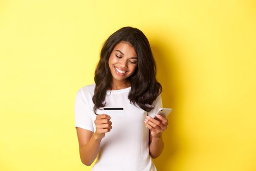 Young attractive woman shopping online, using mobile phone with credit card and smiling, standing over yellow background.