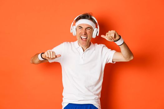 Portrait of handsome middle-aged male athlete, listening music in headphones during fitness training, dancing over orange background.