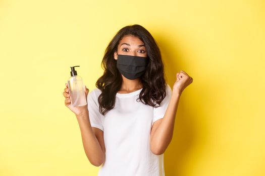 Concept of covid-19, social distancing and lifestyle. Image of happy african-american woman in face mask, feeling happy about founding good hand sanitizer, rejoicing over yellow background.