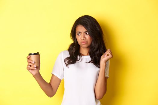 Portrait of african-american girl looking disappointed at cup, dont like bad coffee, standing over yellow background.