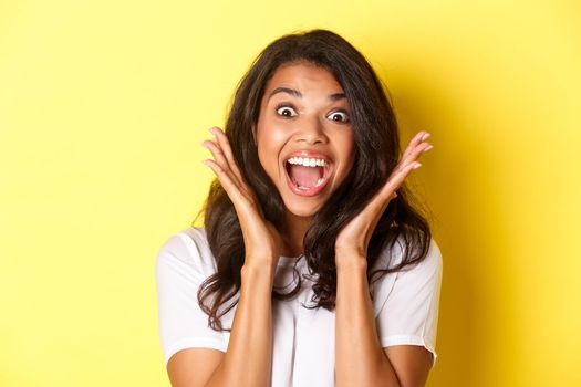 Close-up of excited african-american female model, gasping and looking amazed, raising hands up and seing something cool, standing over yellow background.
