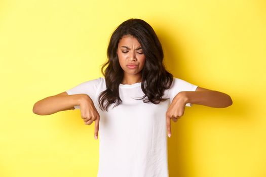 Portrait of sad and disappointed african-american girl, complaining about something bad, looking and pointing fingers down with displeased face, standing over yellow background.