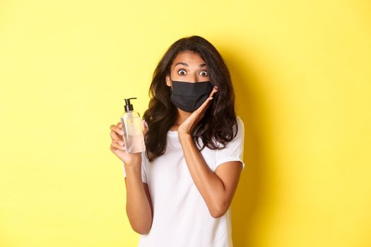 Concept of covid-19, social distancing and lifestyle. Image of impressed african-american girl in face mask, showing good hand sanitizer, recommending product, yellow background.