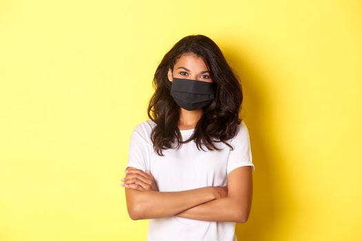 Concept of coronavirus, pandemic and lifestyle. Portrait of young african-american woman in black face mask, smiling and looking confident with hands crossed on chest, yellow background.