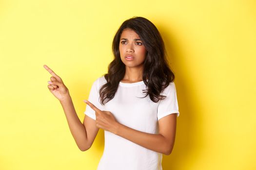 Portrait of doubtful and anxious african-american woman, frowning and pointing fingers at upper left corner, showing something disturbing, standing over yellow background.