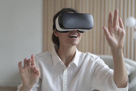 Happy female freelancer wearing VR headset while working remotely online from home, raising hands up trying to touch objects in virtual reality. Girl sits at desk and interacts with 3d visualization