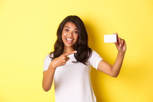 Image of beautiful african-american woman in white t-shirt, pointing at credit card and smiling, recommending bank, standing over yellow background.