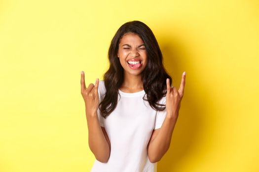 Image of excited and carefree african-american girl, enjoying something cool, making rock-on signs and smiling, standing over yellow background.