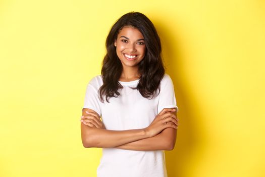 Image of happy smiling african-american girl in white t-shirt, cross arms on chest and looking confident, standing over yellow background.