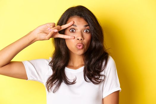 Image of silly and cute teenage african-american girl, showing peace sign and pouting for kiss, standing over yellow background.