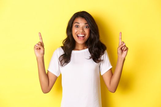 Portrait of amazed, beautiful african-american girl in white t-shirt, reacting to cool advertisement, pointing fingers up, standing over yellow background.