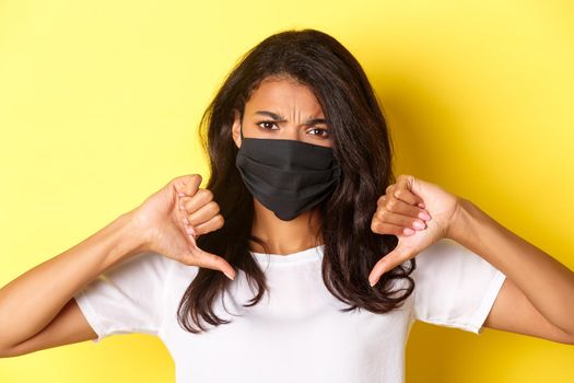 Concept of covid-19, social distancing and lifestyle. Close-up of displeased african-american girl in face mask, frowning upset and showing thumbs-down to disagree, yellow background.