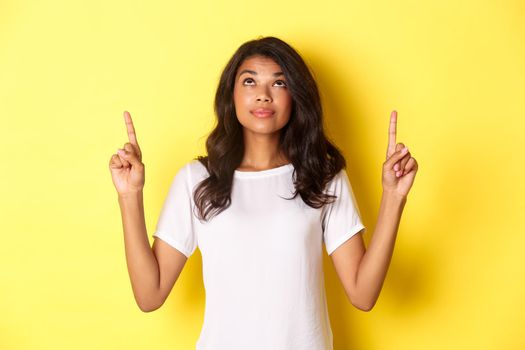 Portrait of attractive african-american female model, wearing white t-shirt, looking and pointing fingers up with curious smile, showing promo, standing over yellow background.