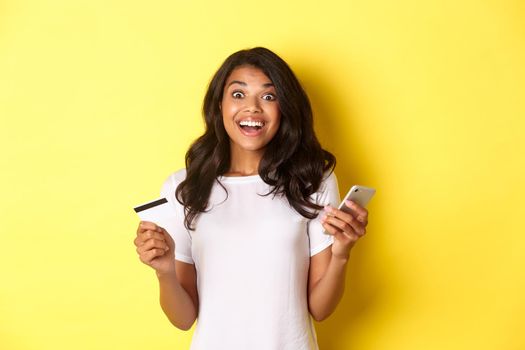 Portrait of amazed african-american girl, holding smartphone and credit card, standing over yellow background.