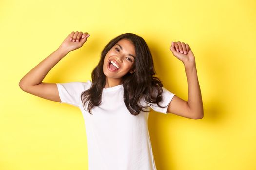 Image of cheerful african-american girl winning, looking happy and celebrating victory, triumphing about achievement, standing over yellow background.