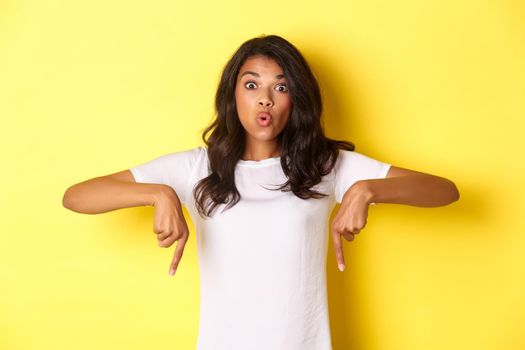 Portrait of attractive african-american girl, pointing fingers down and looking amazed, showing something curious, standing over yellow background.