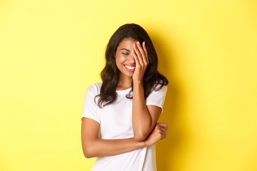 Portrait of beautiful and happy african-american girl, silly laughing and covering her face with hand, standing over yellow background.