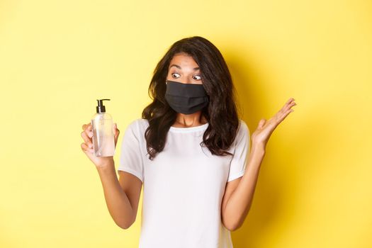 Concept of covid-19, social distancing and lifestyle. Image of amazed african-american girl, wearing face mask, looking at hand sanitizer excited, standing over yellow background.
