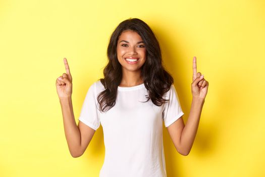 Portrait of attractive and confident african-american female model, wearing white t-shirt, pointing fingers up at promo logo, standing over yellow background.