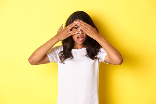 Portrait of embarrassed and shocked african-american girl, shut her eyes but peeking through fingers at something disturbing, standing over yellow background.
