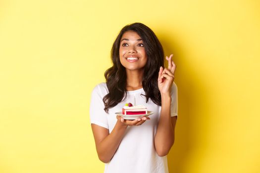 Image of cute and dreamy african-american girl, crossing fingers, holding birthday cake and looking left while making a wish, celebrating b-day over yellow background.