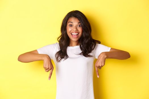 Portrait of amused african-american girl showing announcement, smiling and pointing fingers down at happy news, standing over yellow background.