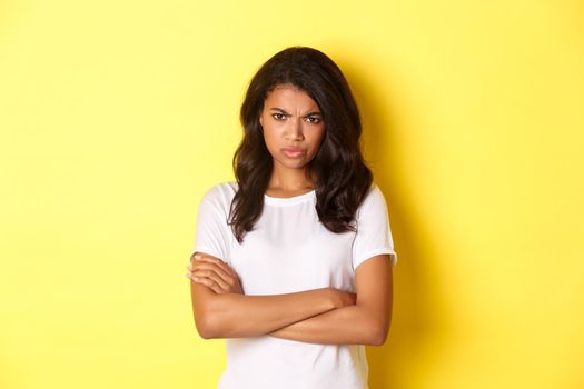 Image of cute african-american girl feeling angry, cross arms on chest and frowning, mad at you, standing over yellow background.