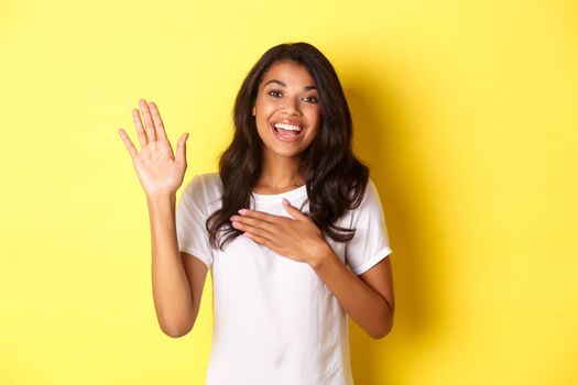 Portrait of cute and sincere african-american girl, making a promise, holding on hand raised and another on heart, swearing tell truth, standing over yellow background.