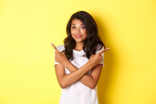 Portrait of cute indecisive african american girl, pointing fingers sideways and shrugging, asking advice with choices, standing over yellow background.