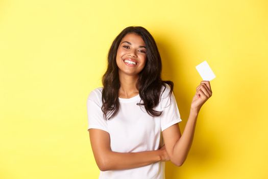 Portrait of beautiful african-american woman in white t-shirt, smiling pleased and showing credit card for shopping, standing over yellow background.