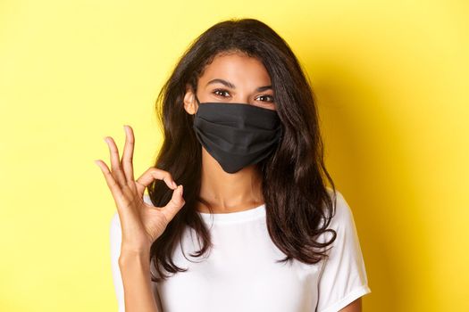 Concept of coronavirus, pandemic and lifestyle. Close-up of pretty african-american woman in black face mask, showing okay sign in approval, praise good work, yellow background.