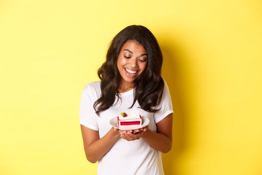 Portrait of attractive african-american woman, looking at delicious piece of cake and smiling, standing over yellow background.