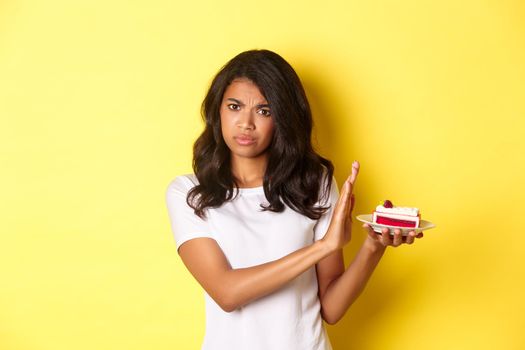 Image of displeased african-american girl decline to eat a cake, standing over yellow background.