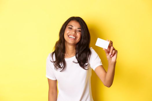 Image of lovely african-american woman smiling happy, showing credit card, standing over yellow background.