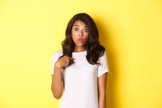 Portrait of surprised african-american girl, pointing finger at herself, standing over yellow background.