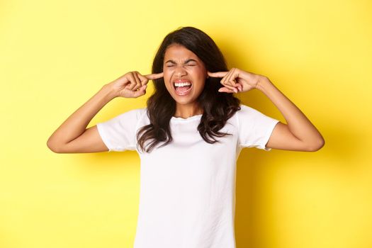 Image of pissed-off african-american girl, cant stand loud bothering noise, shut ears and screaming annoyed, standing over yellow background.