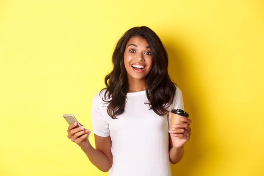 Portrait of attractive african-american girl smiling, holding coffee cup and smartphone, standing over yellow background.