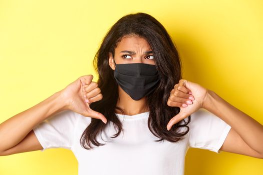 Concept of covid-19, social distancing and lifestyle. Close-up of reluctant and displeased african-american girl, wearing black face mask, showing thumbs-down and frowning upset.